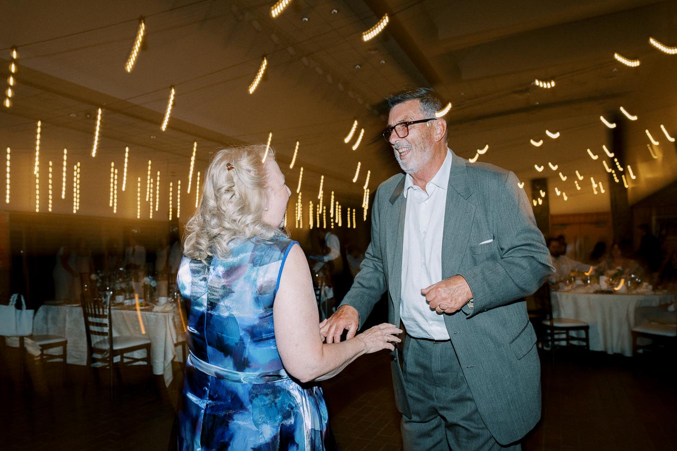 Elderly couple dancing joyfully at a wedding reception with string lights creating a warm ambiance in the background.