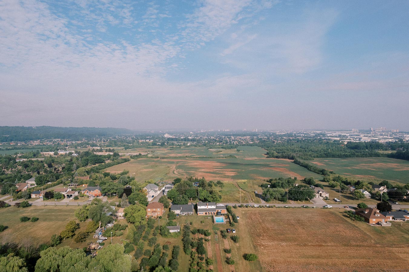 Aerial view of a rural landscape with houses, lush green fields, and trees under a partly cloudy blue sky, showcasing a small town in the distance.