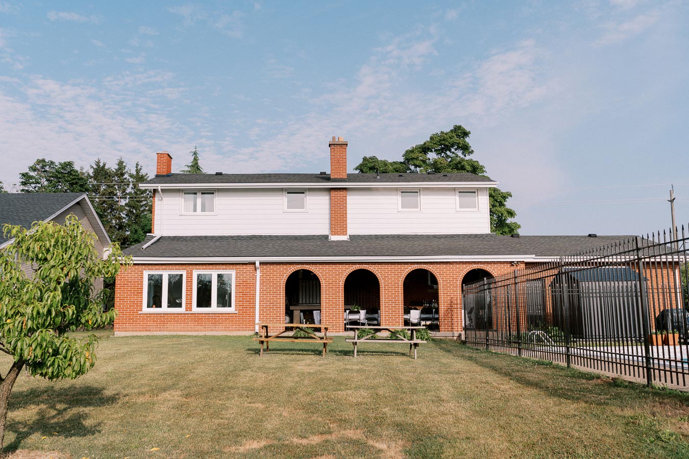 Two-story brick house with arched patio, surrounded by a well-kept lawn and iron fence, under a clear blue sky.