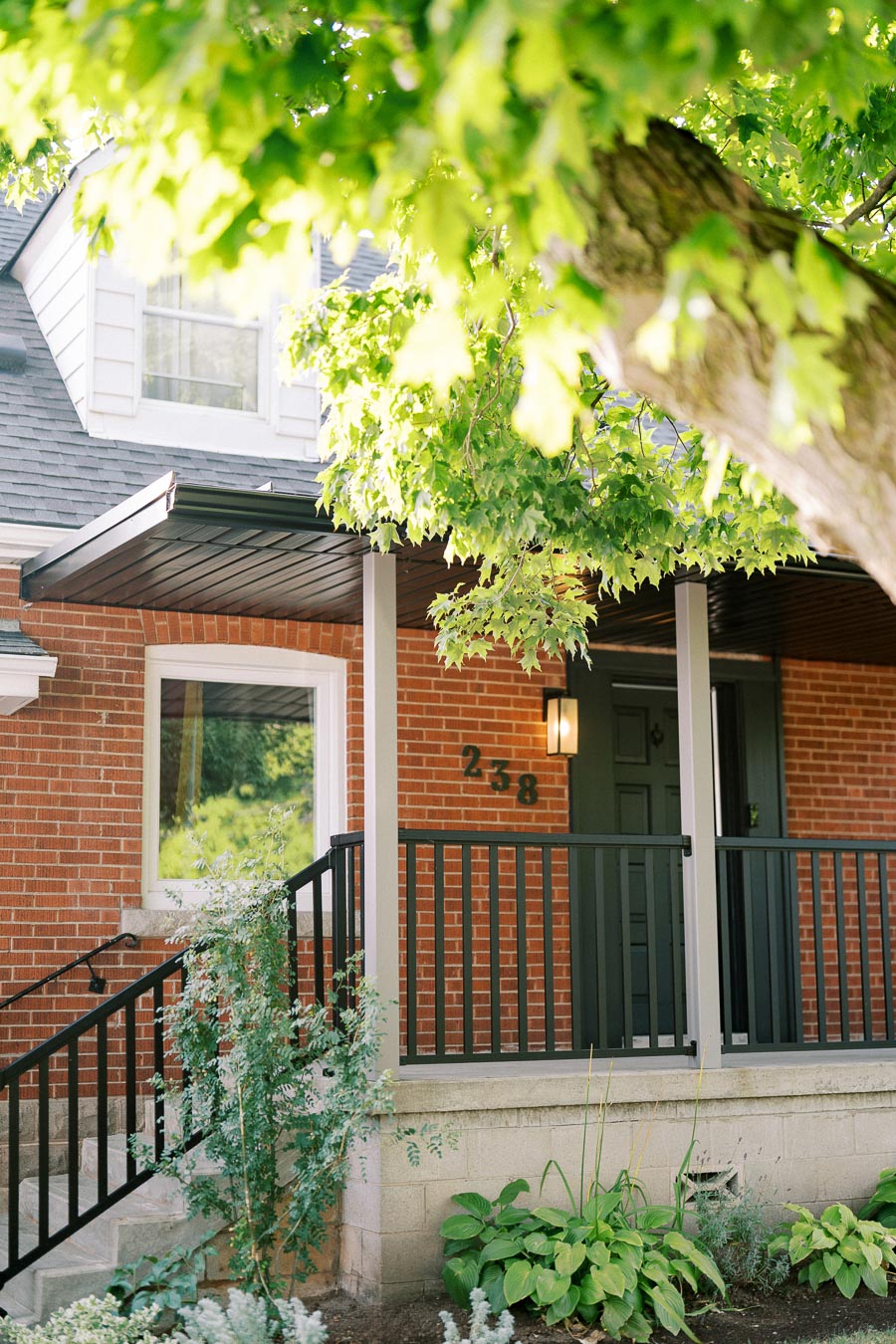 Front view of a charming brick house with a shaded porch adorned by lush green foliage. The house features a prominent black railing and a well-maintained garden, creating a welcoming entrance.