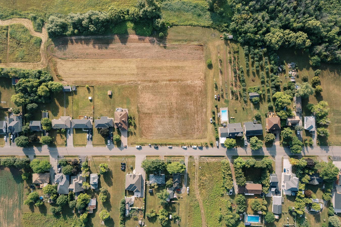 Aerial view of a suburban neighborhood with a central road dividing residential homes and green fields, showcasing a mix of houses, trees, and farmland, under a clear sky.