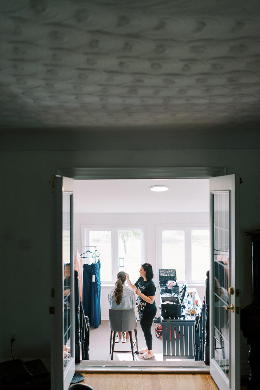 A person receiving makeup application in a bright room with dresses hanging nearby, viewed through glass doors, creating a cozy and intimate atmosphere.