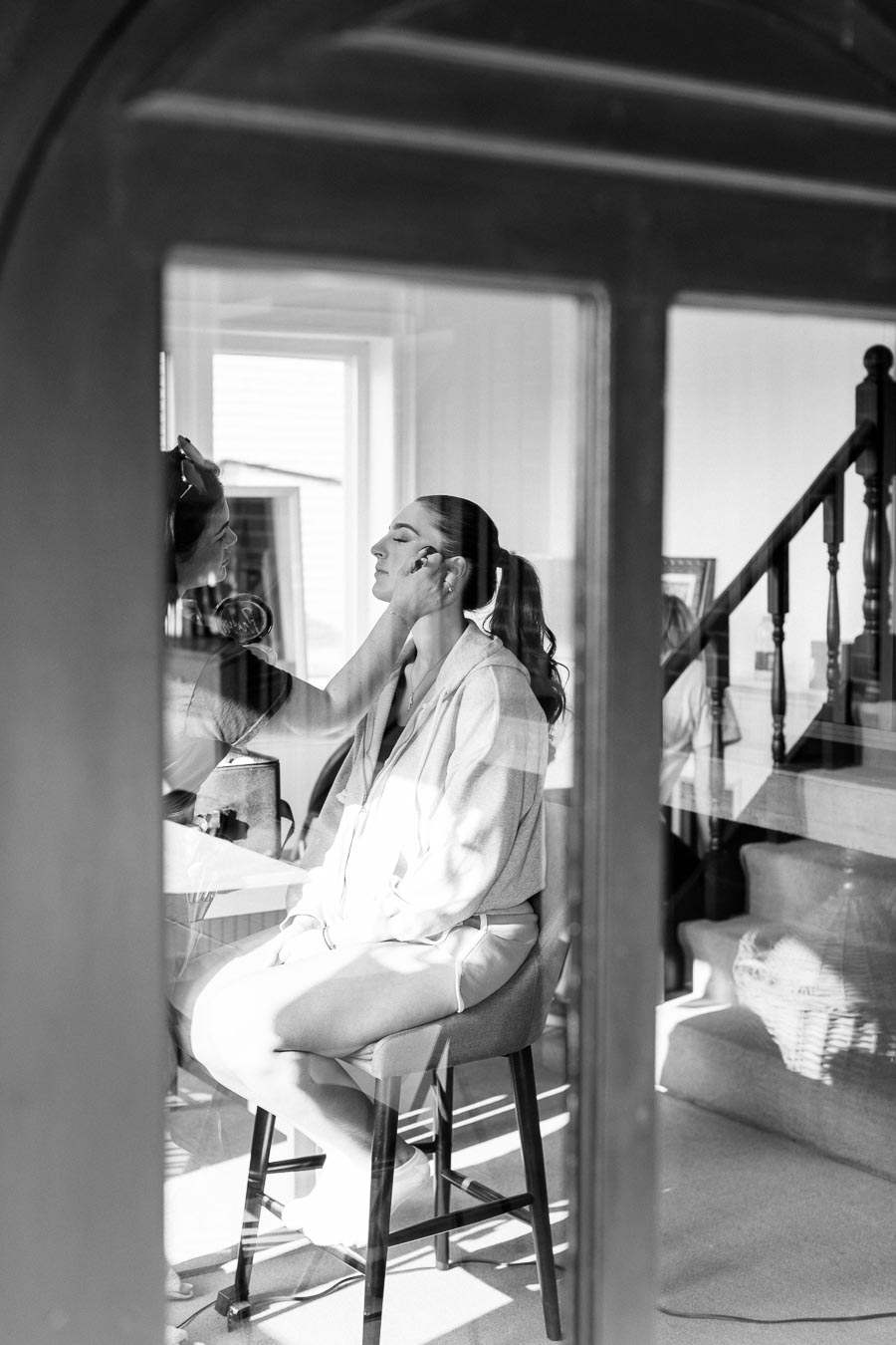 Black and white photo of a woman getting her makeup done, sitting on a stool in a well-lit home setting, with stairs in the background visible through a window.