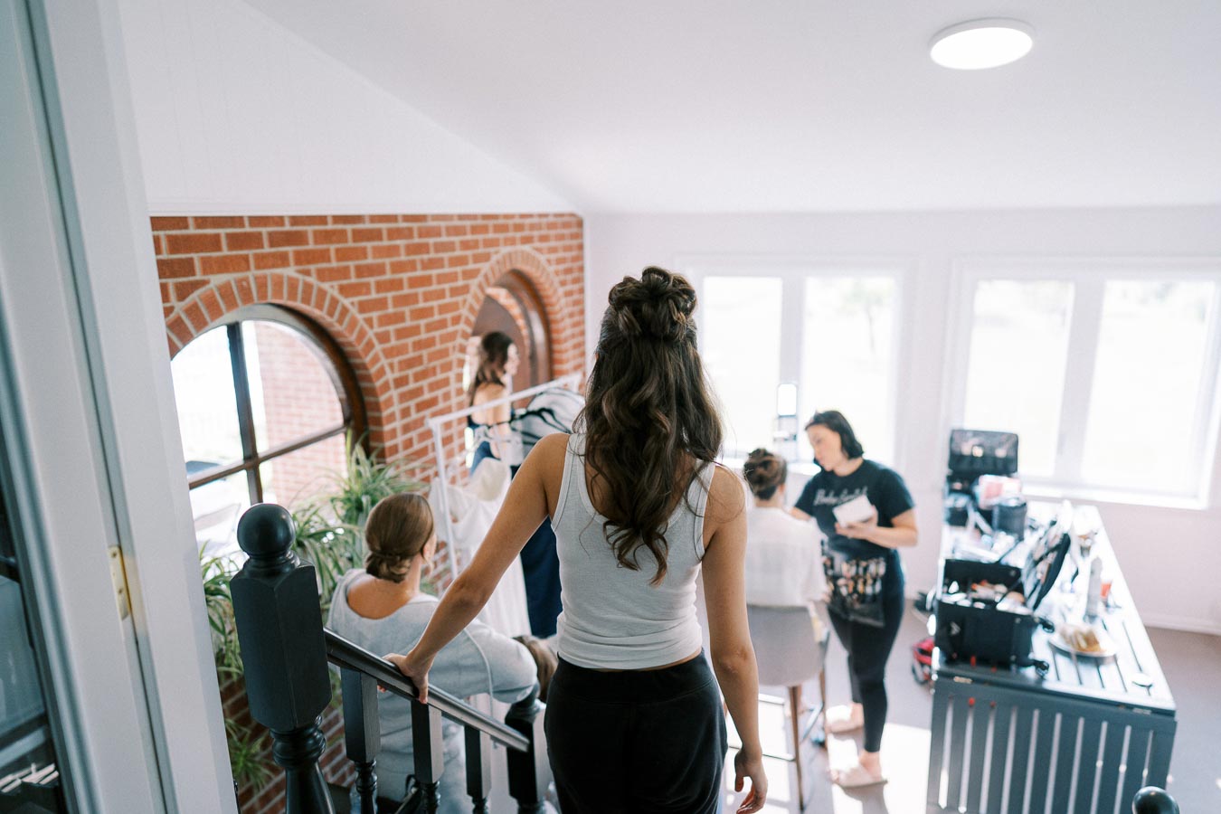 A group of people preparing for an event in a bright room with brick accents, featuring a woman descending stairs in the foreground and a makeup artist attending to a client in the background.