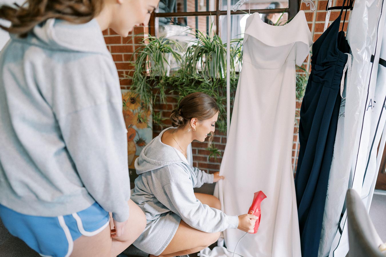 Two young women preparing for a wedding, one using a steamer on a white bridal gown while the other watches.
