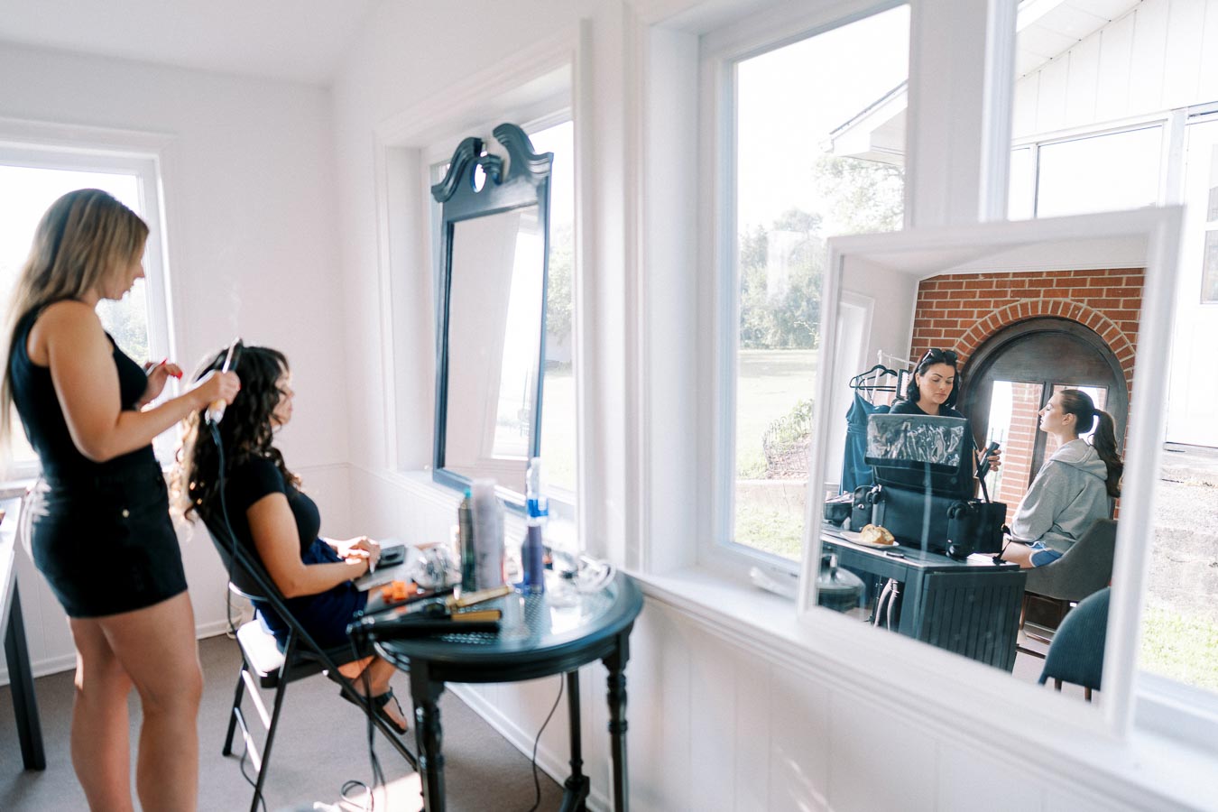 Two women are engaged in hairstyling in a bright salon. One woman is curling another's hair inside, while a makeup session is happening outside. The room has large windows, creating a clean, modern atmosphere with natural light. Hairdressing tools and products are visible on a table.