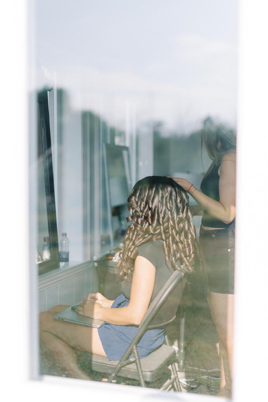 A woman with curled hair sitting in a chair indoors, receiving a hair treatment from a stylist.