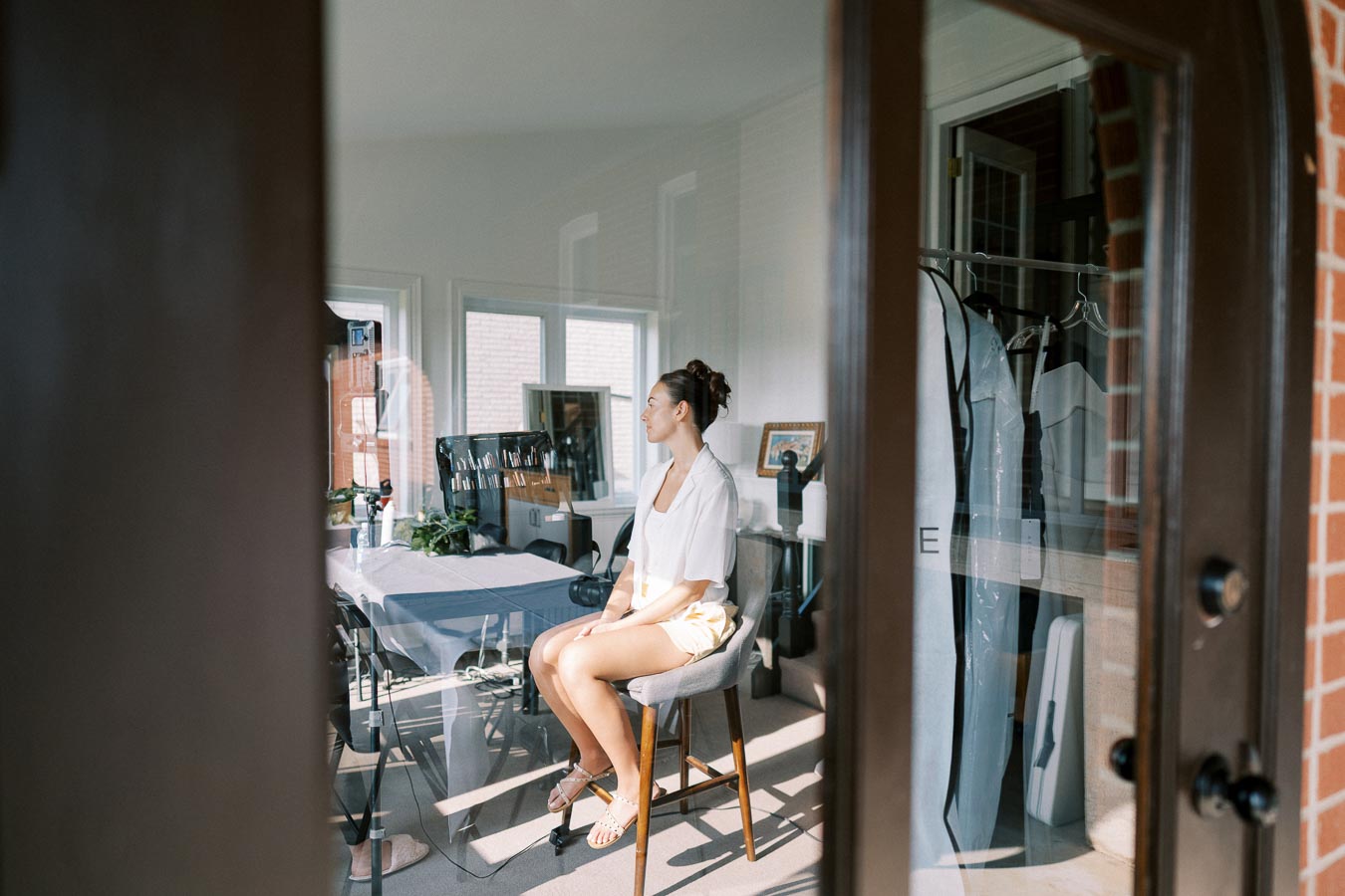 A woman sitting in a sunlit room, with a reflection on the glass door. She is on a high stool, wearing a casual white shirt and shorts. The interior features hanging garments and a table with various items. Bright natural light fills the space, creating a peaceful ambiance.