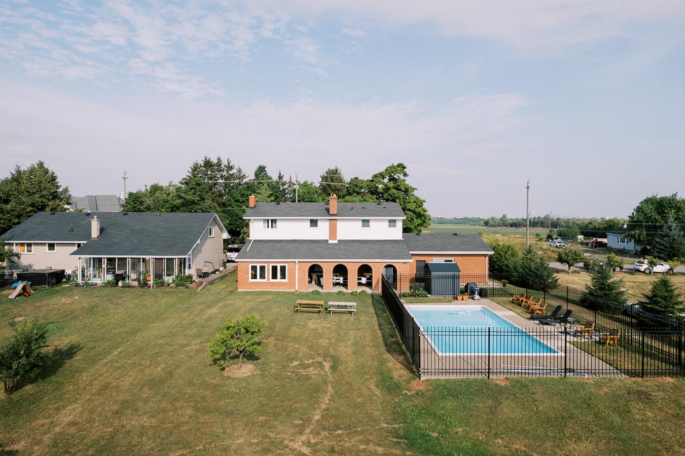 Aerial view of a suburban house with a spacious backyard featuring a lawn, two picnic tables, and a fenced-in swimming pool. The property is surrounded by trees and open space, under a partly cloudy sky.