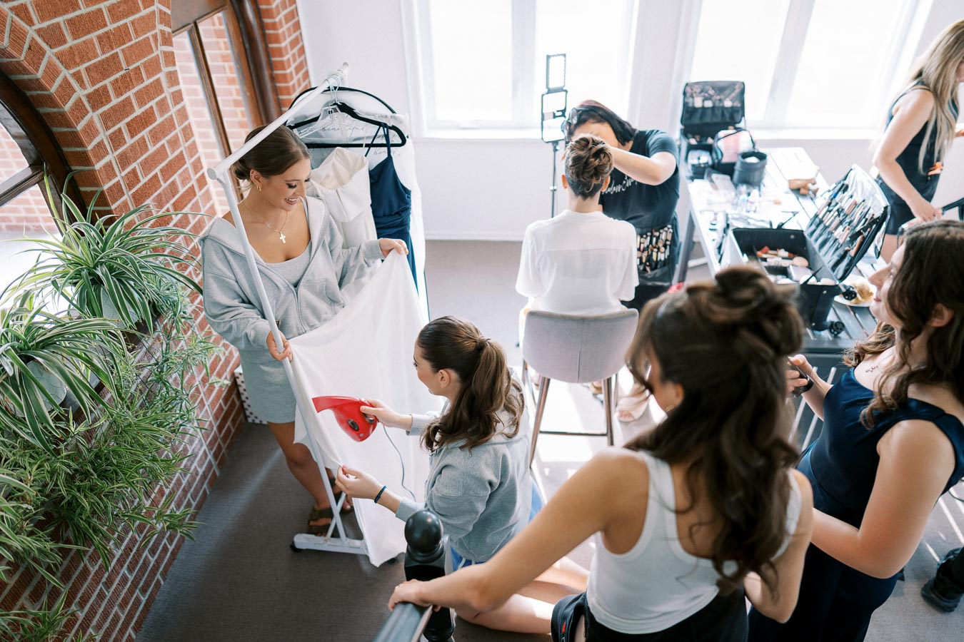 A group of women preparing for an event indoors, with one person steaming a piece of white fabric. Another woman is having her hair styled by a makeup artist, surrounded by beauty tools and attire. The setting is bright with natural light from large windows, enhancing the cheerful and busy atmosphere.