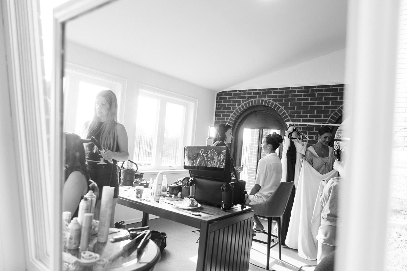 Bridal party getting ready in a bright room, with a stylist curling hair in the foreground and a bride preparing for a wedding in the background, showcasing makeup and hairstyling preparations.
