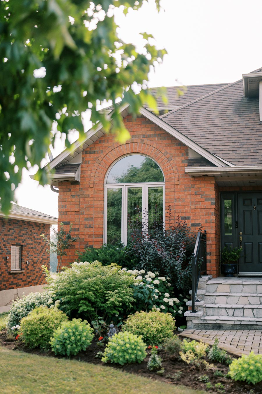 Charming brick house with arched window and lush garden landscape featuring various green shrubs and flowering plants, alongside stone steps leading to a black door.
