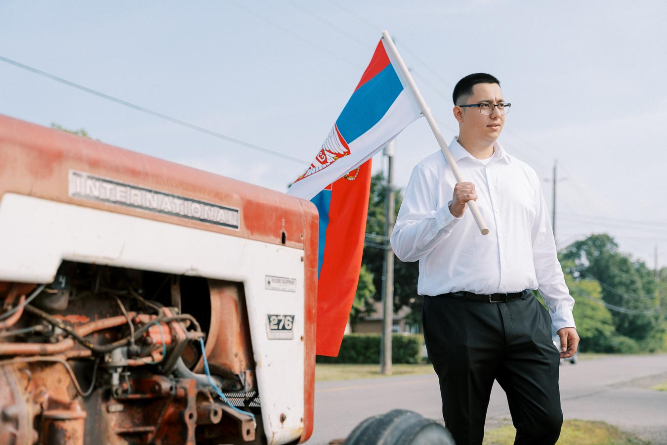 Man standing next to a vintage International tractor, holding a Serbian flag, wearing a white shirt and black pants, set against a rural outdoor backdrop.