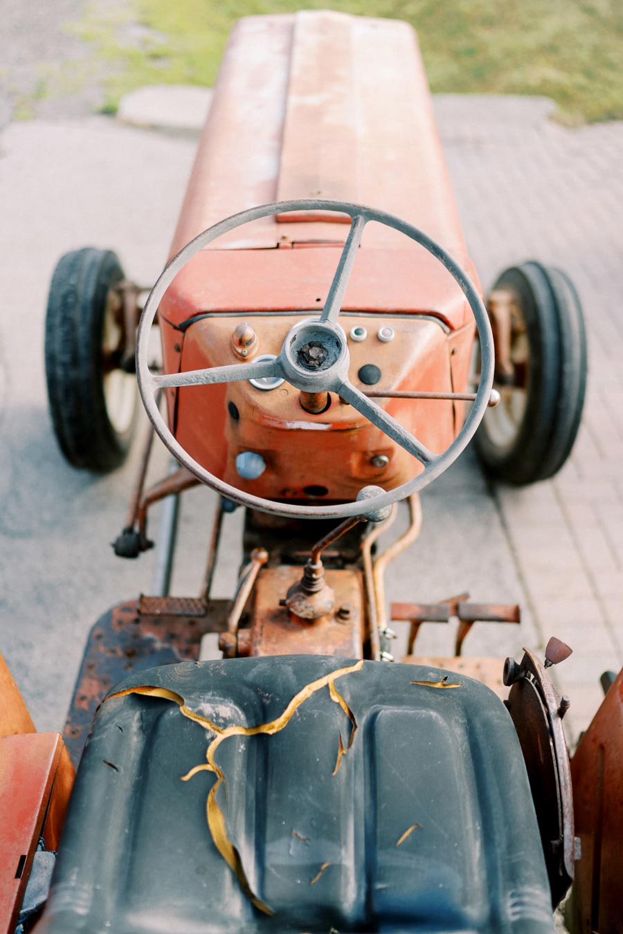 Top view of a vintage red tractor with a weathered steering wheel and controls, parked on a paved surface.