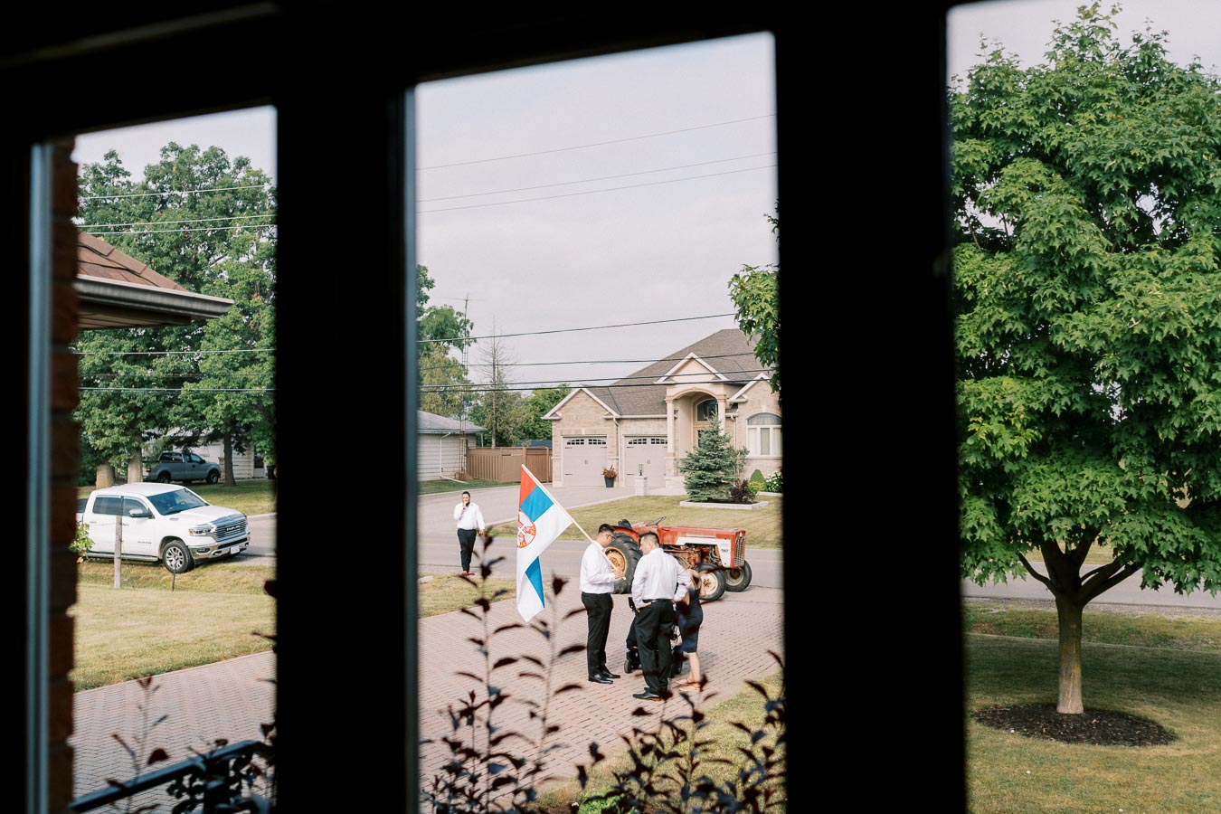 View through a window of people gathered on a driveway, with a red tractor, holding a flag featuring blue, red, and white colors, near a suburban home surrounded by trees and a parked white vehicle. The scene captures a casual outdoor gathering in a residential neighborhood.