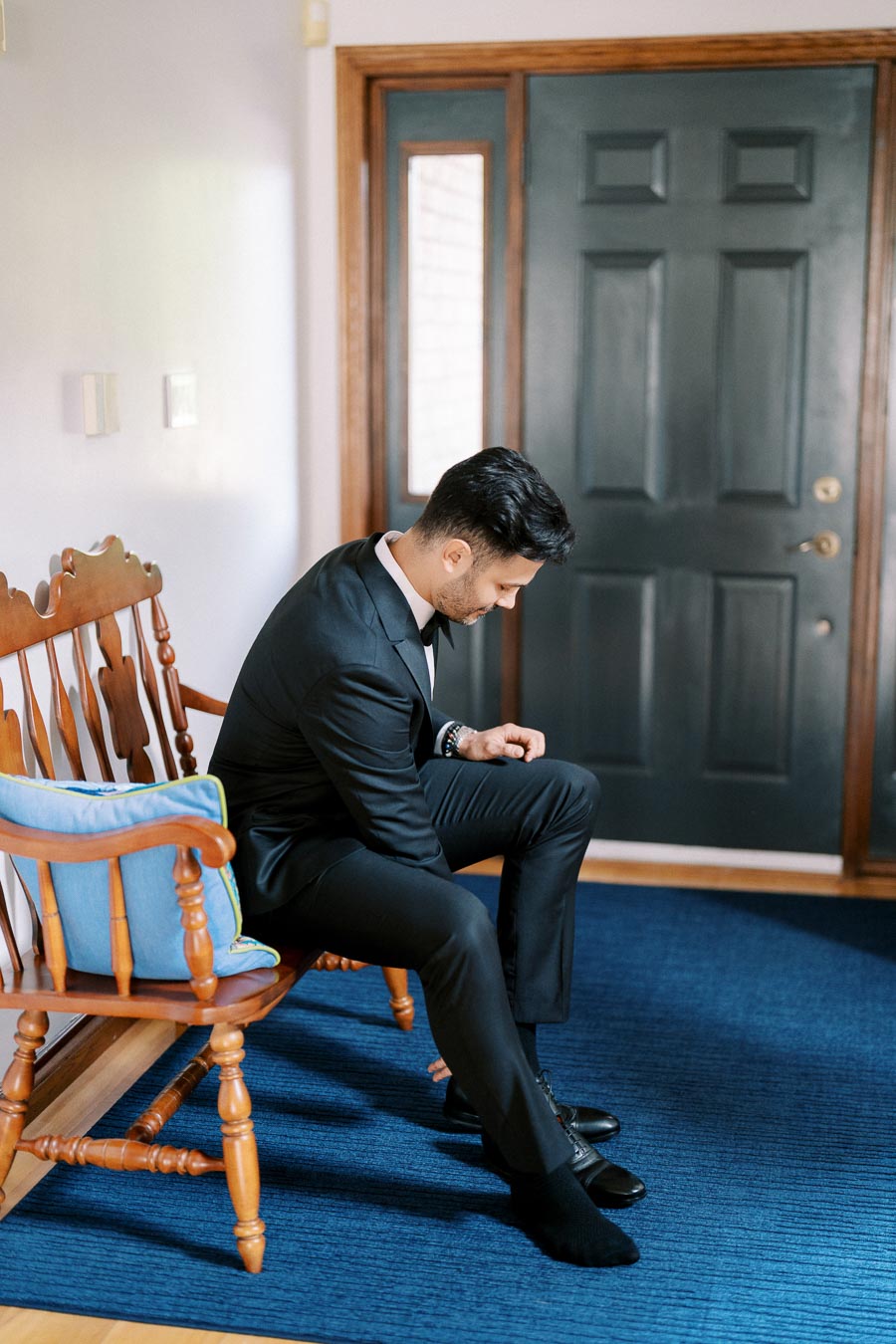A person in a black suit sitting on a wooden bench with a blue cushion, adjusting their shoes near a dark door.