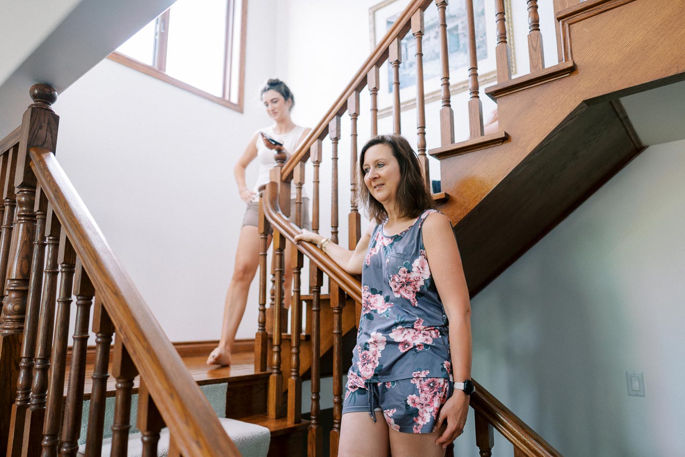 Two women standing on a wooden staircase, one in floral print pajamas, conveying a relaxed and domestic atmosphere in a well-lit home setting.