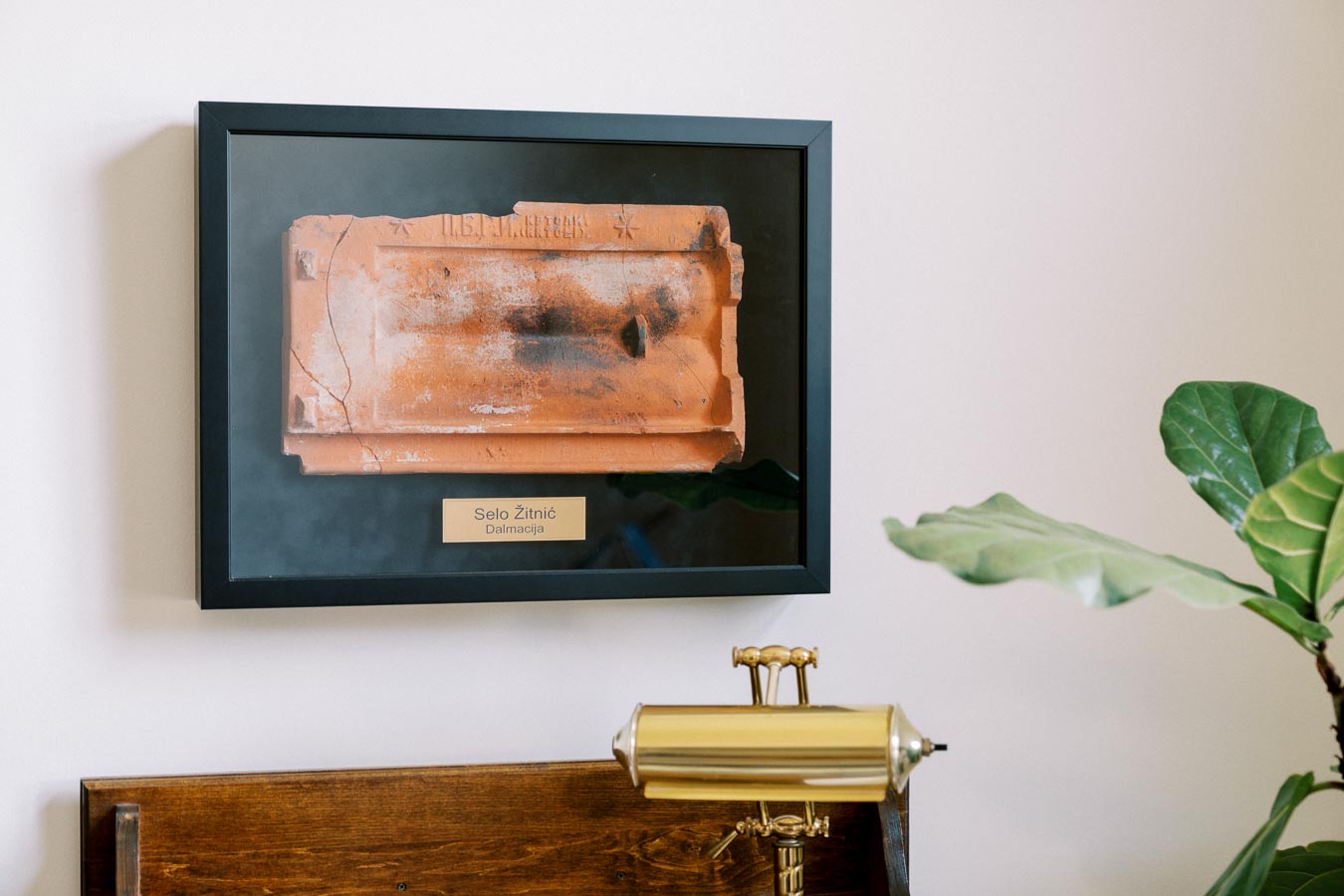 Framed terracotta tile with inscription from Selo Žitnić, Dalmacija, displayed on a wall above a wooden desk with a vintage brass desk lamp and a green plant in the corner.