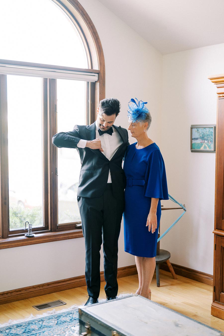 A man in a formal black suit adjusts his bowtie next to an elegantly dressed woman in a blue dress and matching fascinator, standing in a well-lit room with large arched windows.
