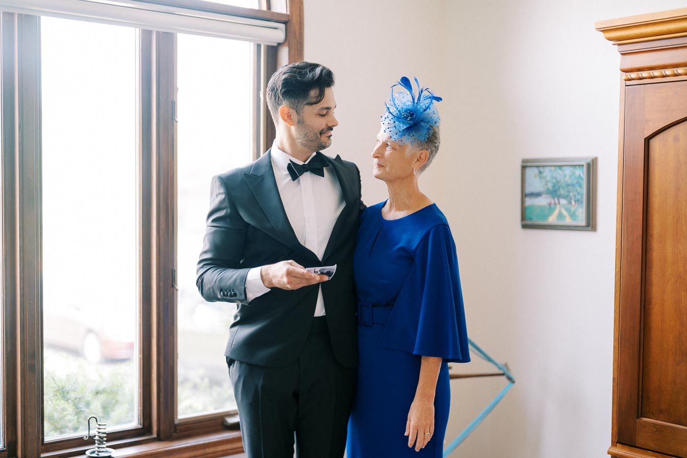 A man in a black tuxedo and a woman in a blue dress with a matching hat stand by a window, looking at each other affectionately.