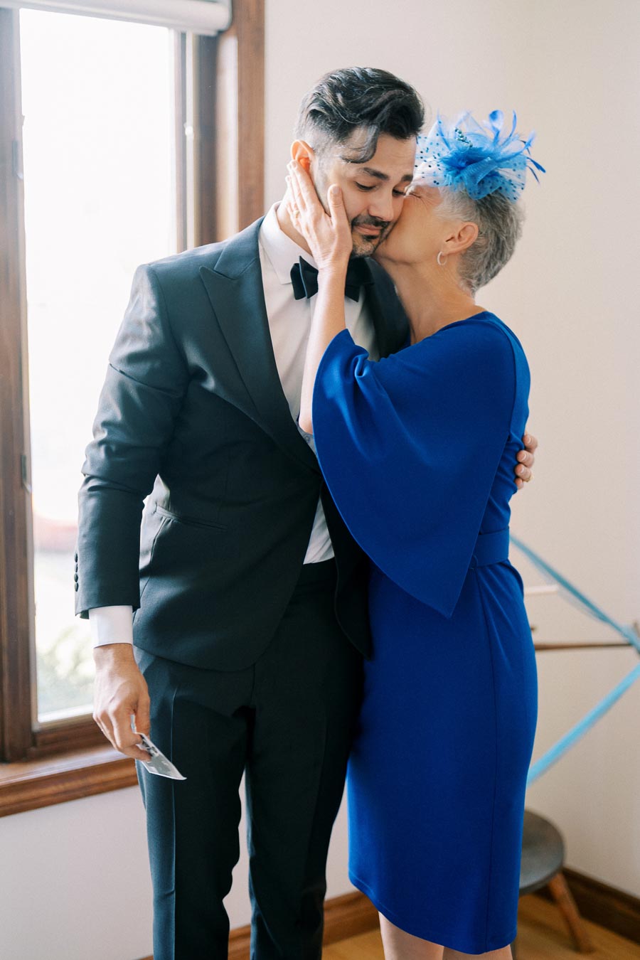 An elegantly dressed woman in a blue dress and fascinator affectionately kisses a man in a black tuxedo, standing by a window with natural light.