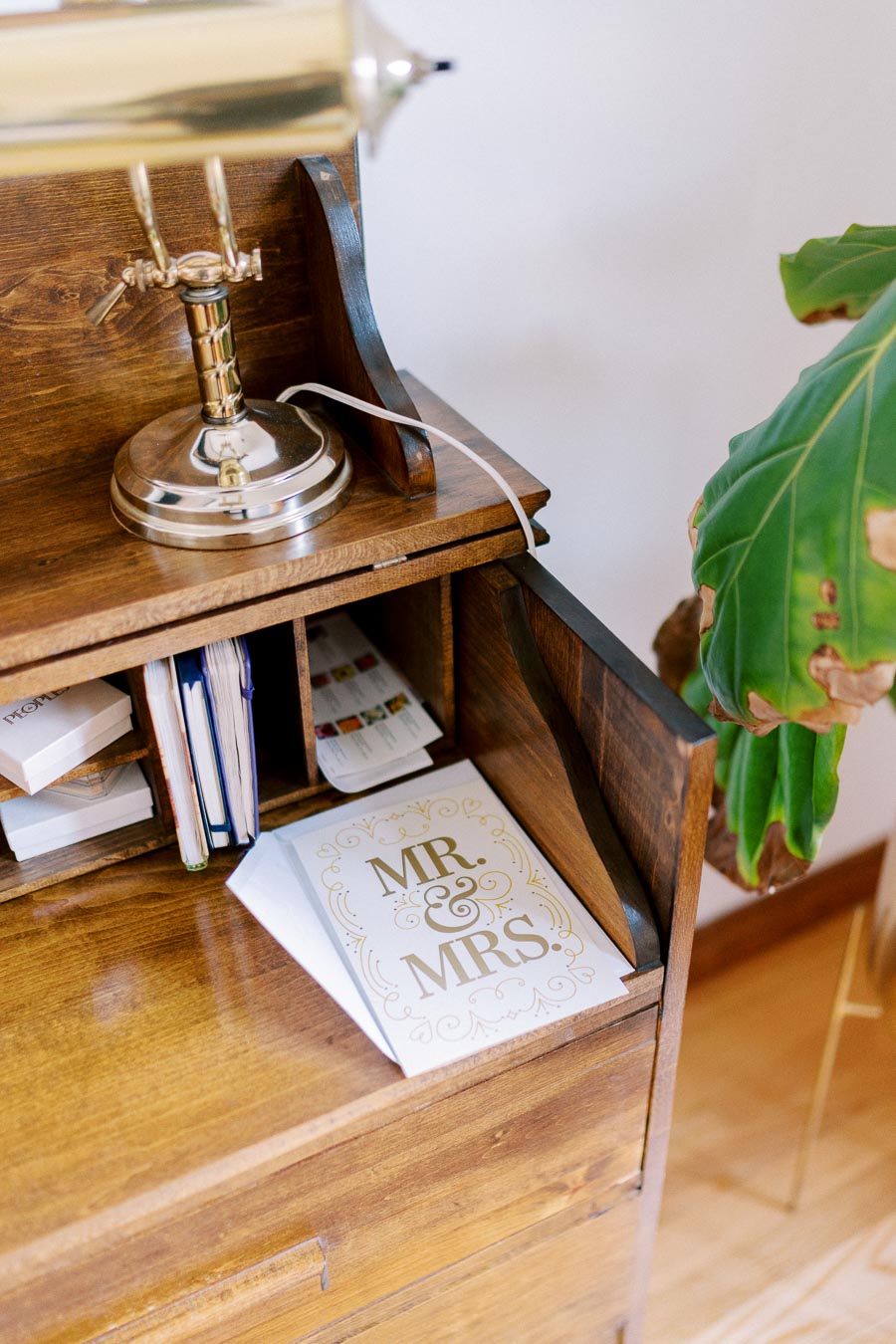 Vintage wooden desk with a brass lamp and a 'Mr. & Mrs.' wedding card, complemented by a green indoor plant for a cozy home office setup.