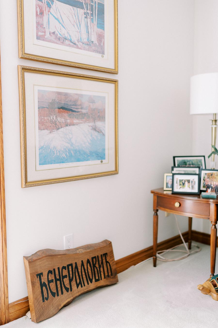 Interior corner featuring framed landscape paintings, a wooden table with family photographs, and a decorative wooden sign with Cyrillic script.