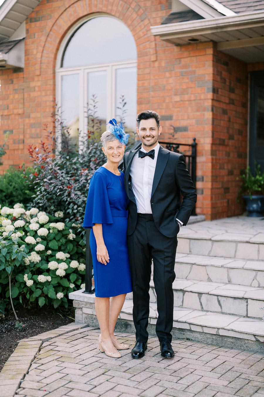 Elegant older woman in a blue dress with a matching fascinator and a young man in a black tuxedo, posing in front of a brick house with lush garden in the background.