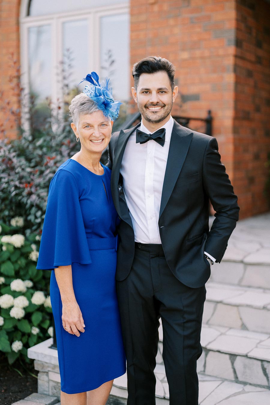 Elegantly dressed older woman in a blue dress with matching fascinator and smiling man in a tuxedo, posing outdoors in front of a brick building.