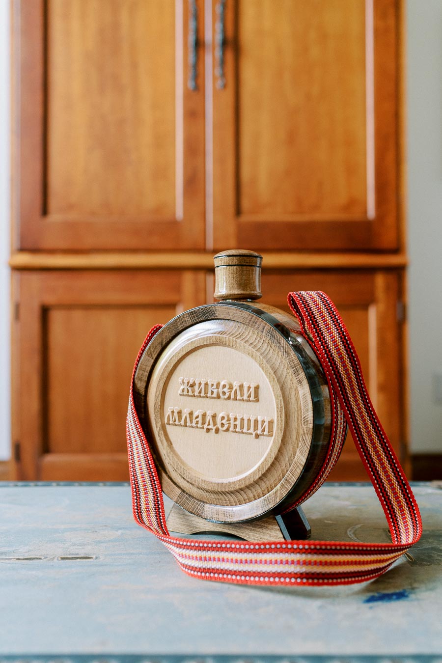 Decorative wooden barrel with traditional embroidery ribbon and Cyrillic text, placed on a rustic wooden table in front of wooden cabinets.