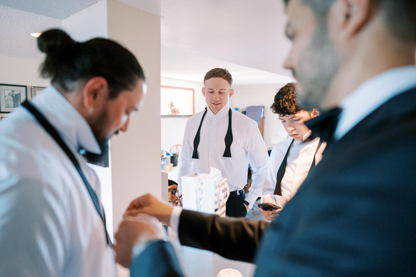 Four men preparing for a formal event, adjusting bow ties and getting dressed in a well-lit room, indicating a pre-wedding or gala gathering.