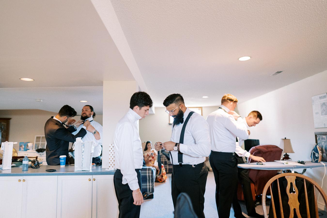 A group of men in formal attire getting ready in a living room, adjusting ties and shirts, with an ironing board and other preparations visible in the background, conveying a wedding or special event scene.