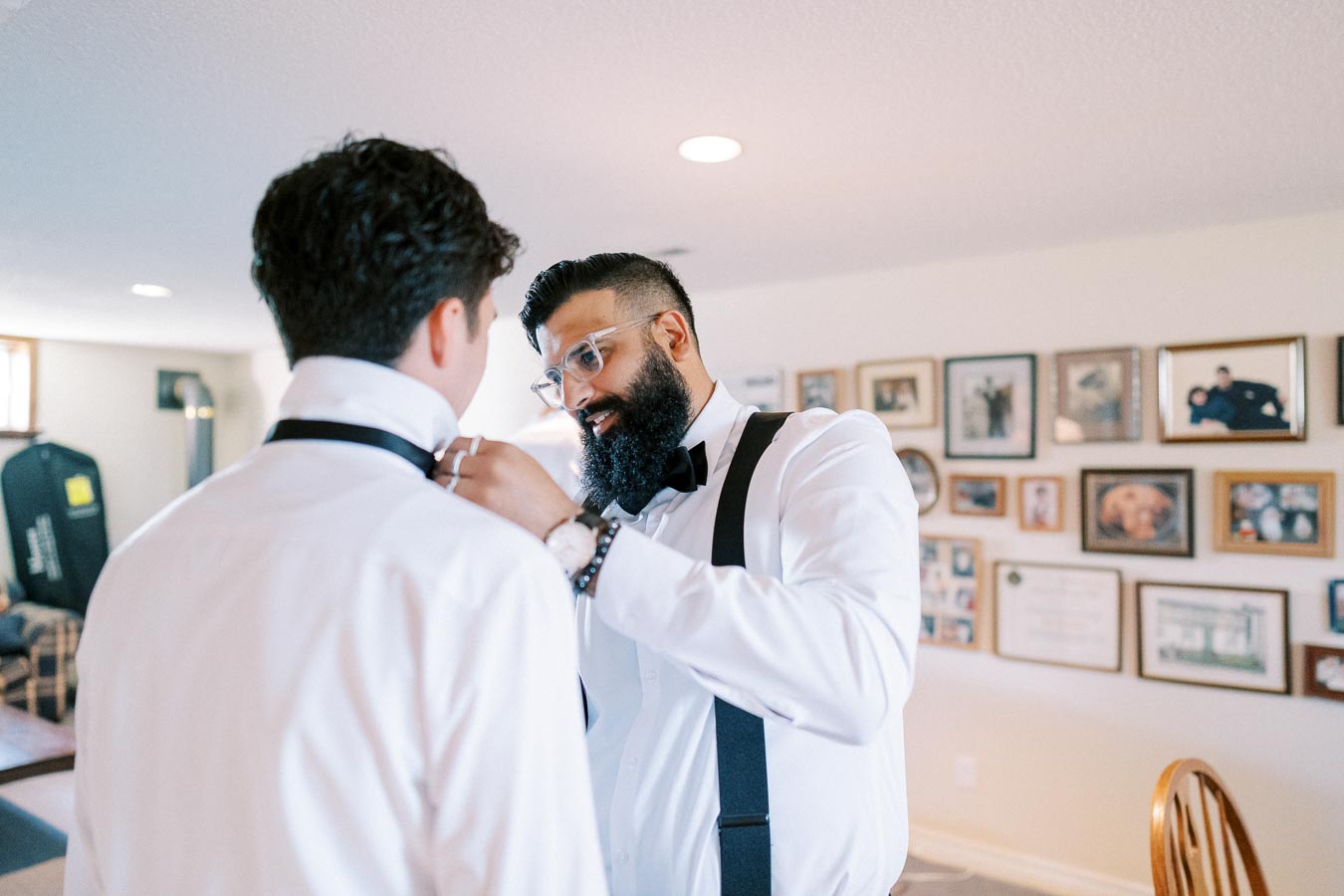 Two men dressed in formal attire, one adjusting the other's bow tie, in a room decorated with numerous framed photos on the wall.