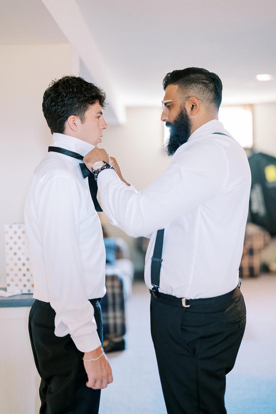 Two men in formal attire preparing for an event, with one helping the other adjust his bow tie in a warmly lit room.