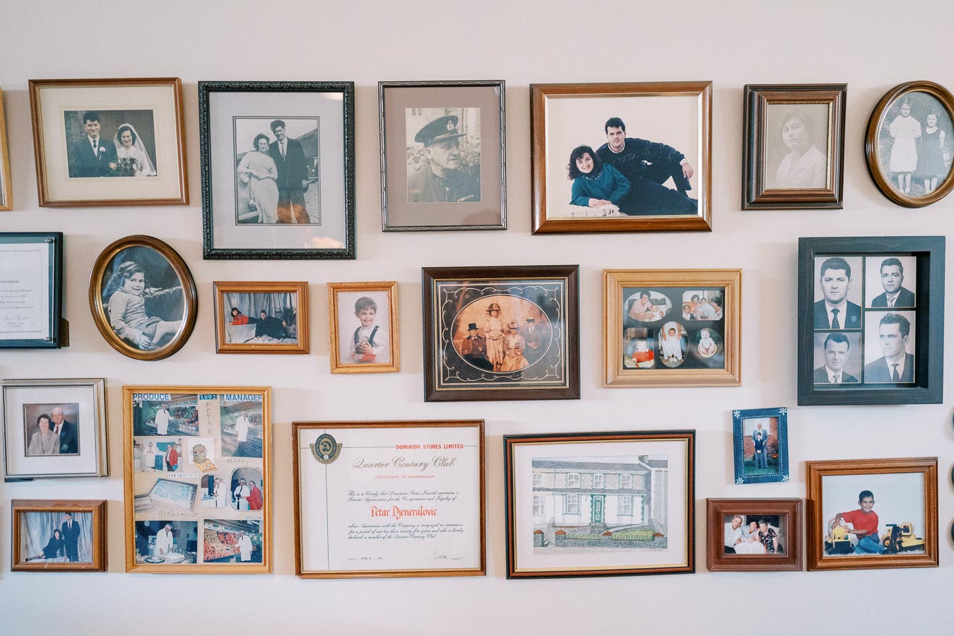 A wall showcasing an assortment of framed family photographs and memorabilia spanning different generations. The display includes a mix of black and white as well as color images. Some frames hold portraits, while others feature certificates and a drawing of a house, creating a diverse and nostalgic family gallery.