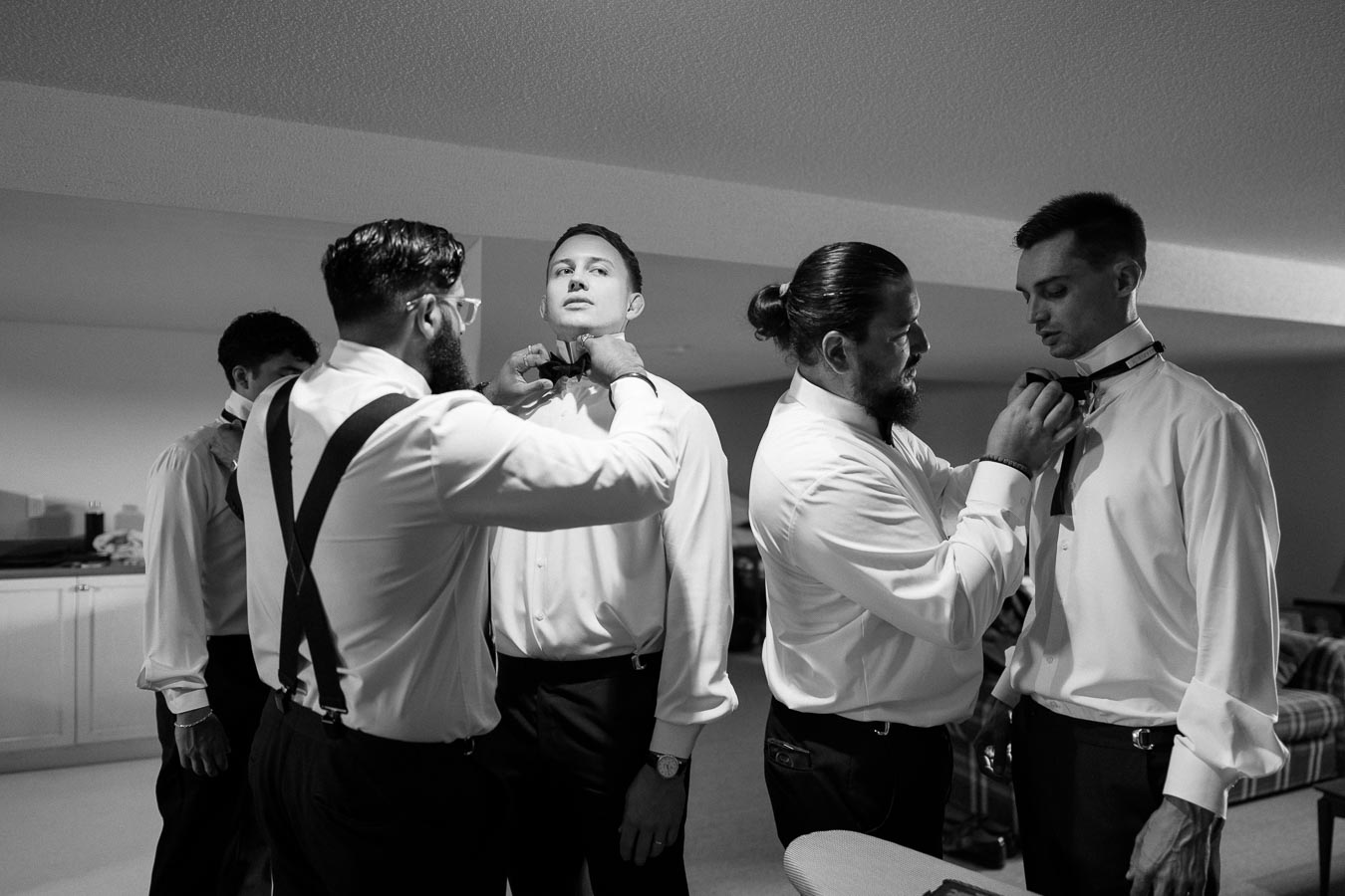 Groomsmen preparing for a wedding, adjusting bow ties and wearing formal suits in a room, showcasing teamwork and preparation.