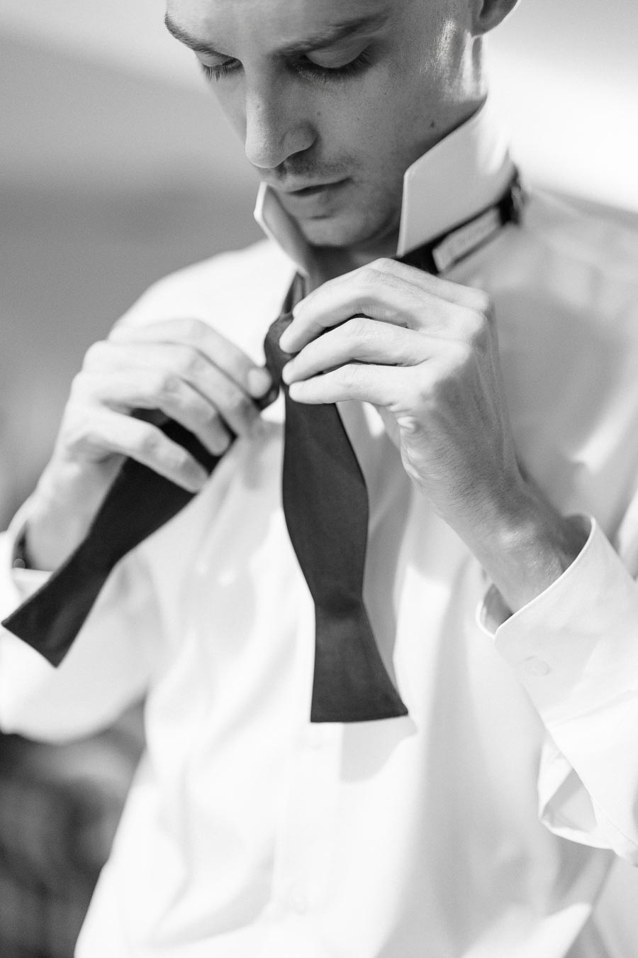 Black and white image of a man tying a bow tie, focusing on the intricate detail of the tie and the hands, capturing a classic style and elegance in formal attire preparation.
