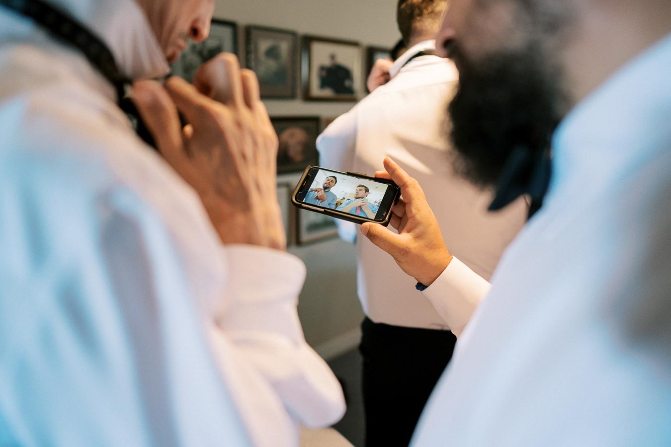 Groom and groomsmen getting ready, adjusting ties while watching a video tutorial on a smartphone before a wedding ceremony.