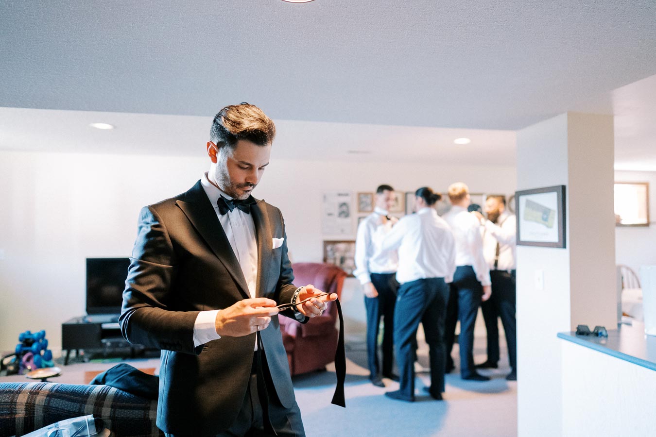 Groom getting ready for wedding in a stylish black tuxedo, adjusting his bow tie in a well-lit room, with groomsmen preparing in background.