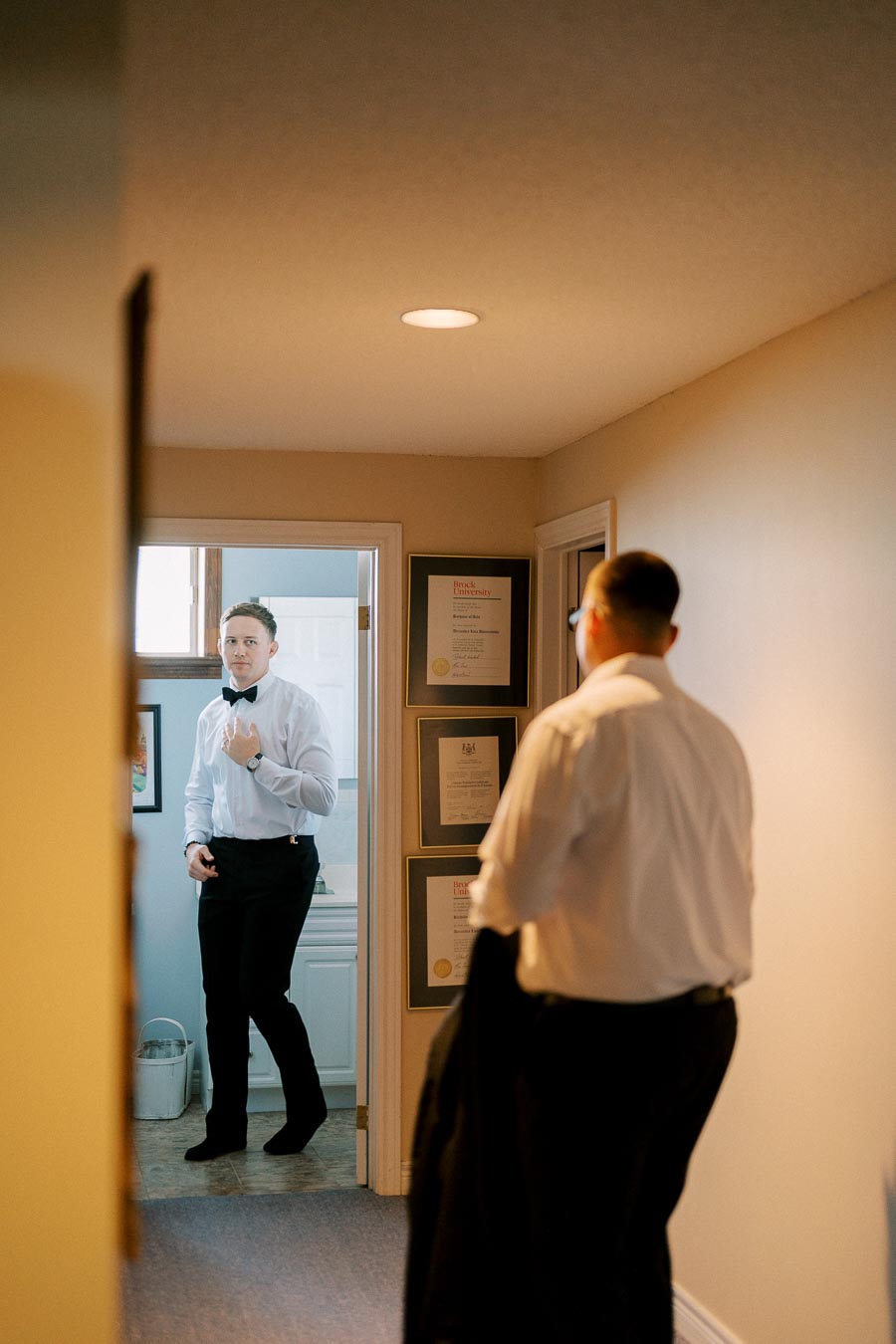 A man in formal attire adjusting his bow tie in a hallway, with framed certificates on the wall, preparing for a formal event.