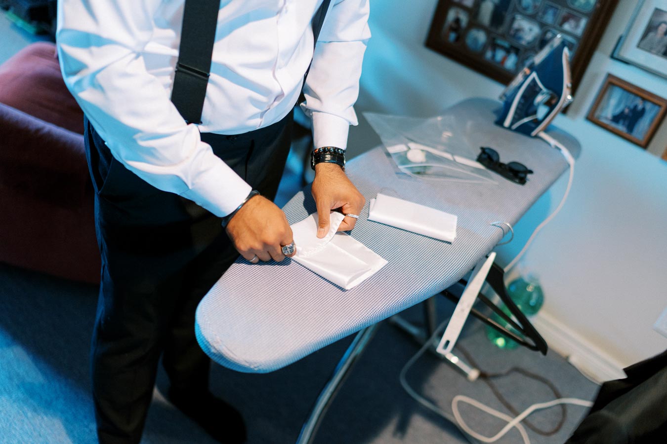Man preparing white pocket square on ironing board with iron and sunglasses nearby, wearing formal white shirt and suspenders.