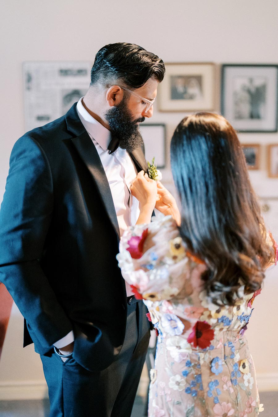A couple preparing for a formal event, with a woman in a floral dress pinning a boutonniere on a man in a black suit and bow tie.