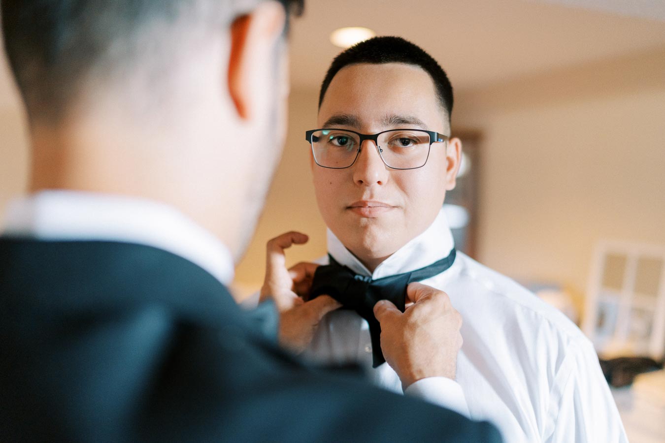 Wedding groom getting ready as an attendant helps adjust his bow tie, showing a close-up of the preparation process in a softly lit room.