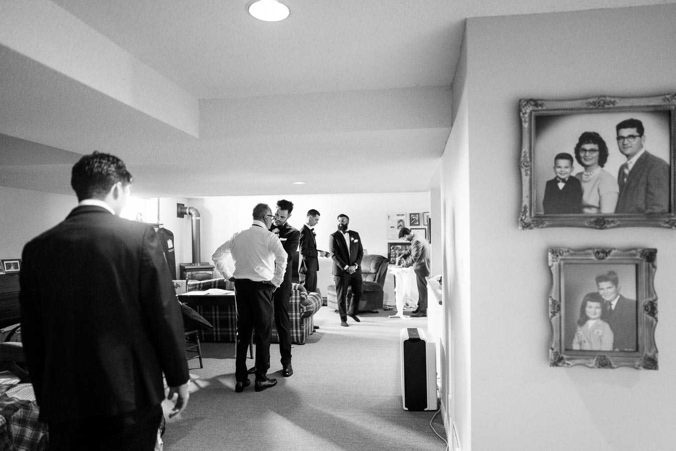 Group of men in suits, preparing for a wedding in a cozy, vintage-style living room with family photos on the wall.