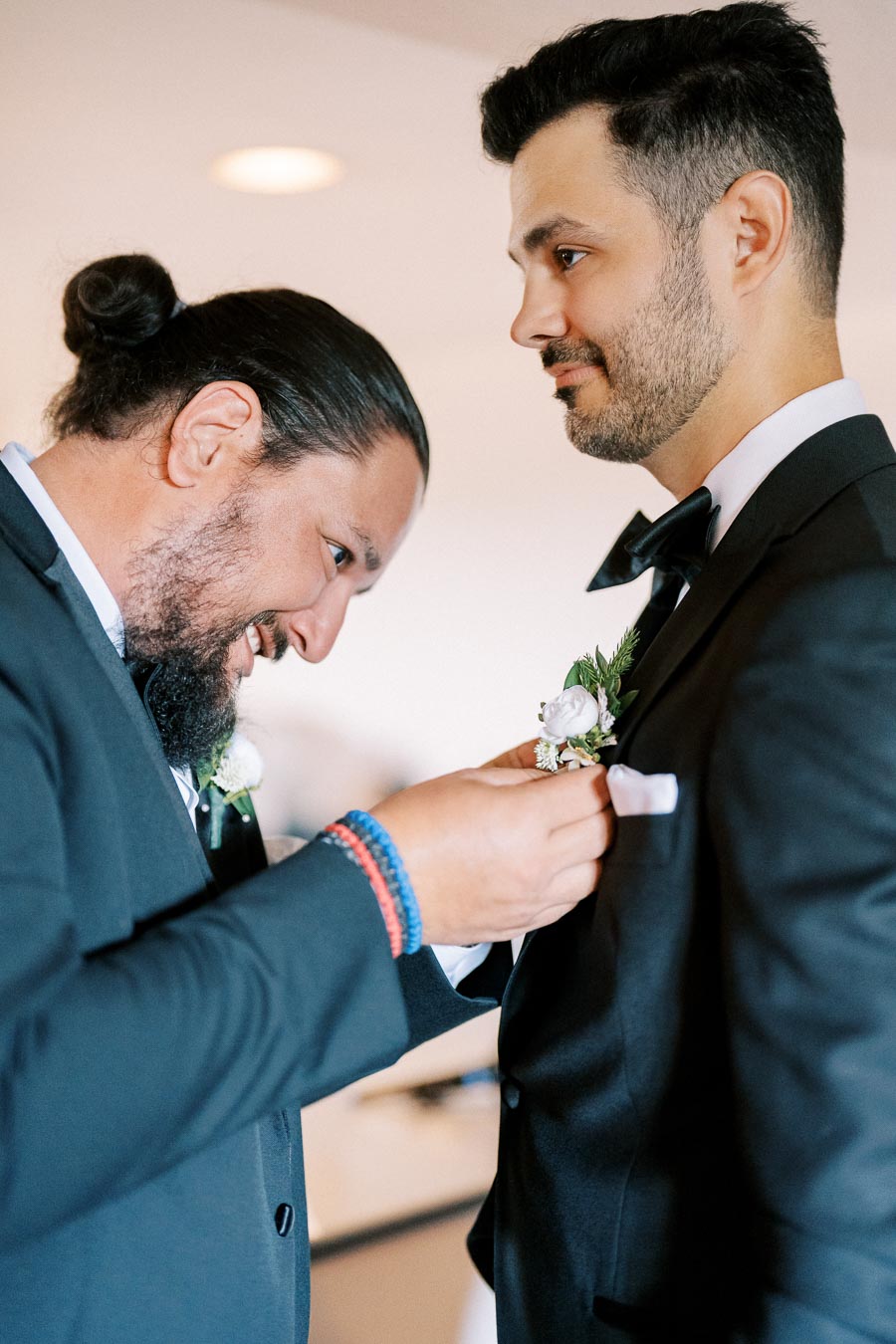 Two men in formal suits, preparing for a wedding. One man is helping adjust a boutonniere on the other man's suit jacket, showcasing friendship and attention to detail. Both are smiling, capturing a joyful wedding preparation moment.