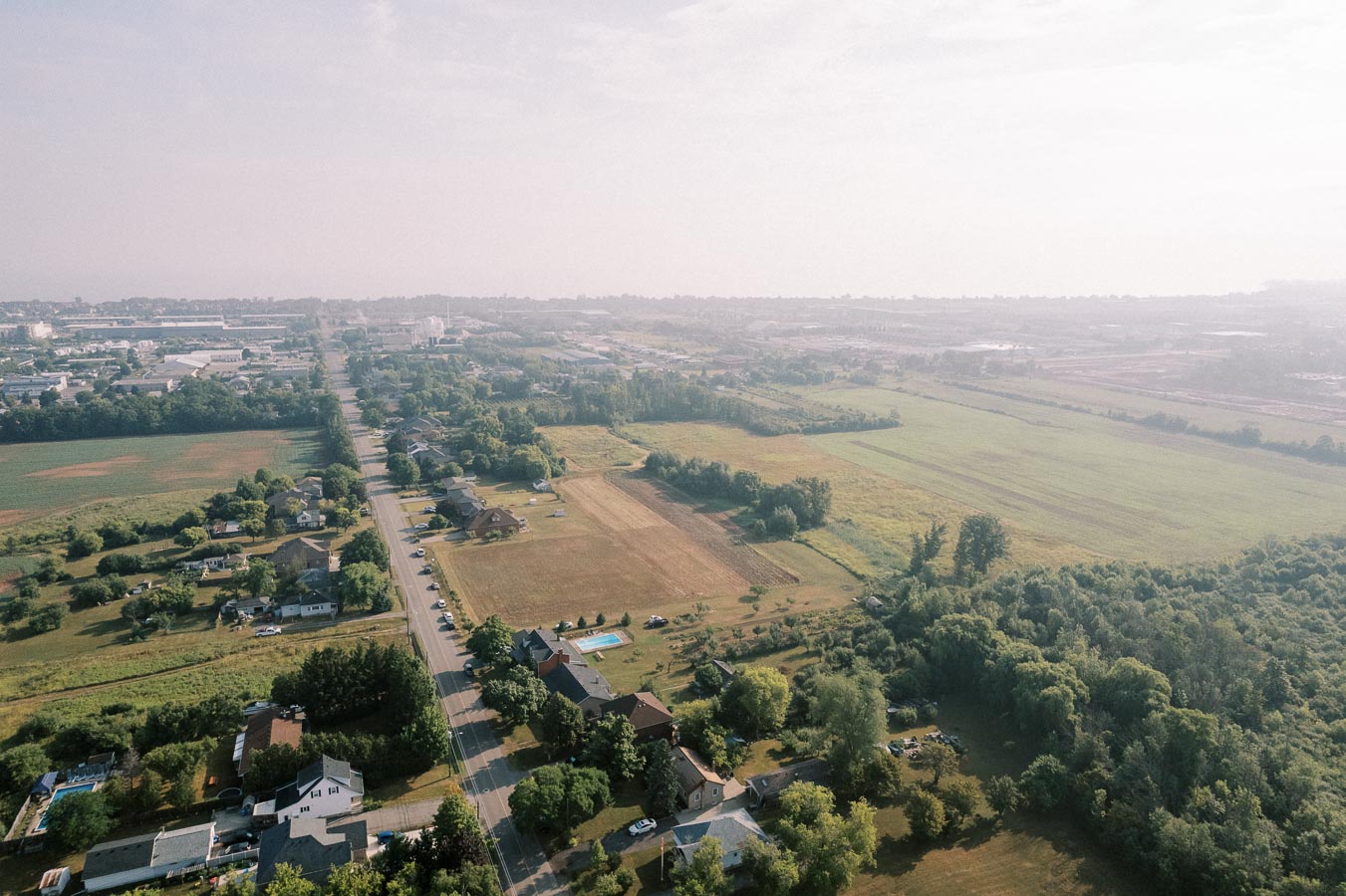 Aerial view of a sprawling rural landscape with green fields, residential houses, and a straight road leading toward the horizon under a clear sky.