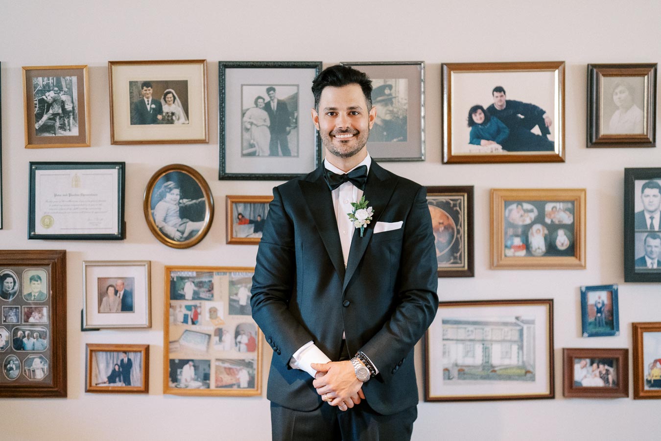 Dapper man in a black tuxedo with bow tie standing in front of a wall of framed family photos.