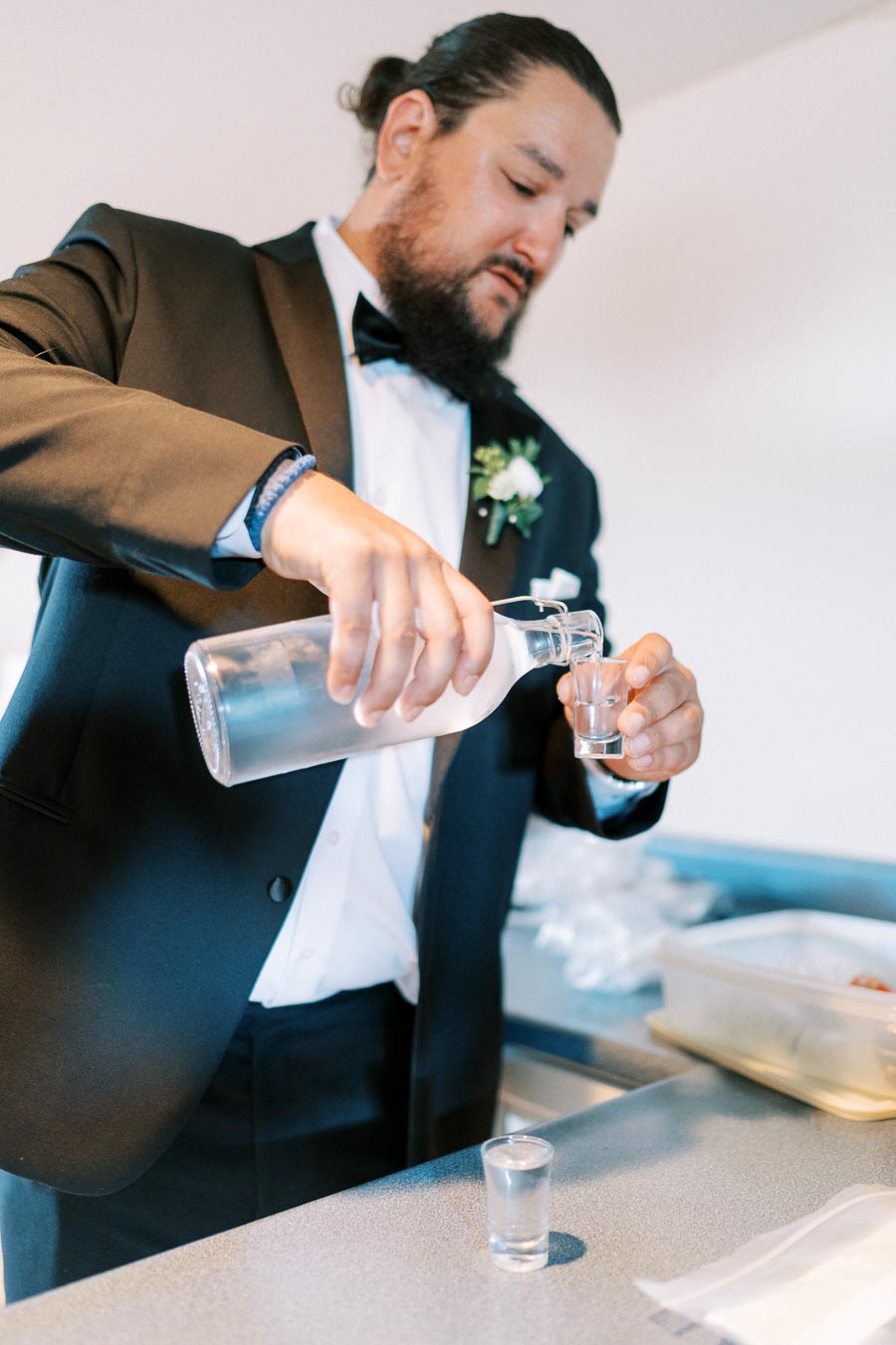 Man in formal attire pouring clear liquid into a shot glass in a kitchen setting, preparing for a celebration or event.