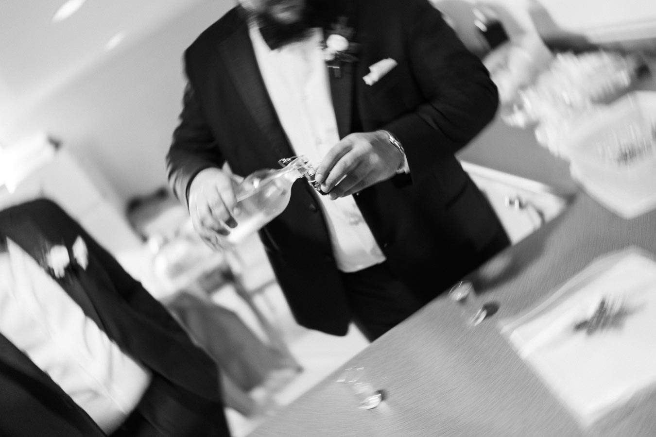 Blurred black and white image of a person in formal attire pouring a drink from a bottle into a glass, possibly at a social event or gathering.