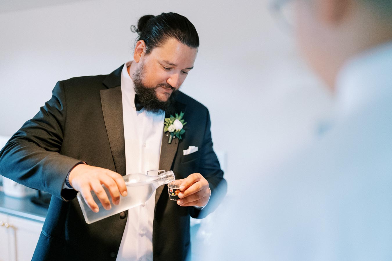 A man in a tuxedo pours a clear beverage into a shot glass, wearing a boutonniere, possibly preparing for a wedding toast.